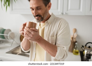 Bearded Man Smiling While Holding Cup Of Warm Tea In Blurred Kitchen