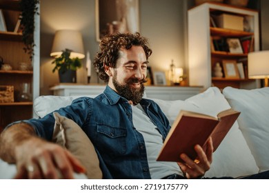 Bearded man smiling while reading a book on a comfortable sofa at home