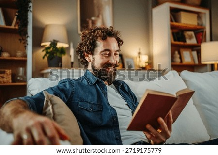 Bearded man smiling while reading a book on a comfortable sofa at home