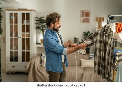 Bearded man smiling while using a handhold garment steamer on a plaid shirt at home, performing casual laundry and self-care in a modern apartment setting