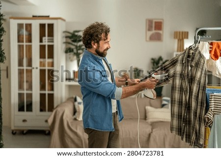 Bearded man smiling while using a handhold garment steamer on a plaid shirt at home, performing casual laundry and self-care in a modern apartment setting
