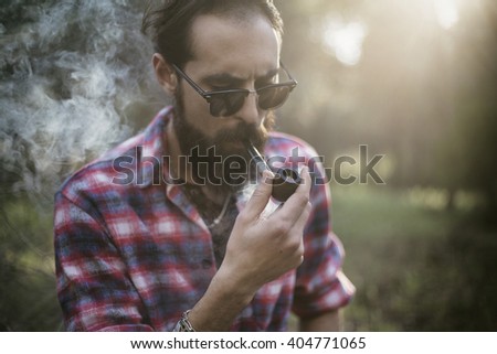 Bearded man smokes a pipe in the wood with beautiful sunbeam