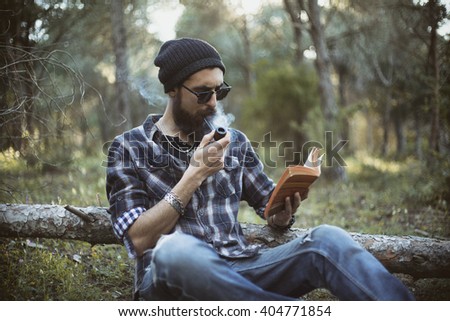 Bearded man smokes a pipe in the wood while reading a book