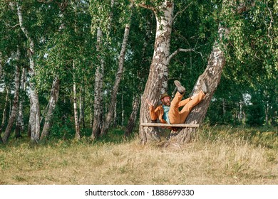 A Bearded Man In A Suit And Hat In The Forest Falls From A Homemade Bench In The Form Of A Pole. Fall In The Woods Badly Sat Down