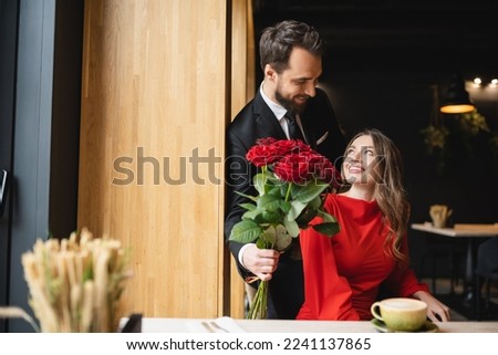 bearded man in suit holding bouquet of red roses near smiling girlfriend on valentines day