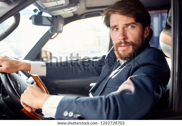 A bearded man in a suit sits behind the wheel of\
a car and looks away        