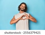 Bearded man in a tank top, blue backdrop smiling and showing a heart shape with hands.