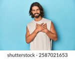 Bearded man in a tank top, blue backdrop has friendly expression, pressing palm to chest. Love concept.