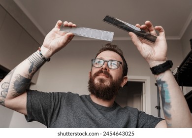 A bearded man with tattoos holds up and examines two strips of film negatives, comparing their details in a well-lit room. - Powered by Shutterstock