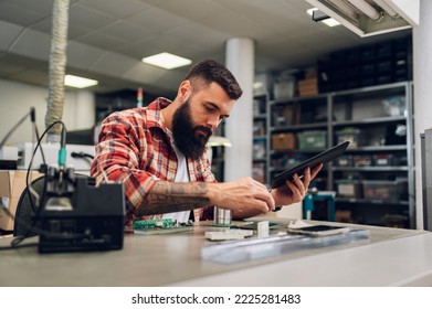 Bearded man technician testing computer part in electronics workshop. Engineer using digital tablet and disassembling the parts of a hardware component in a repair shop.