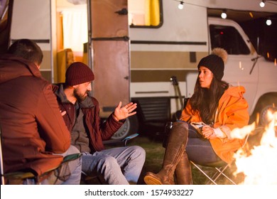 Bearded man telling a story to his friends around camp fire in a cold night of autumn in the mountains. Retro camper van.