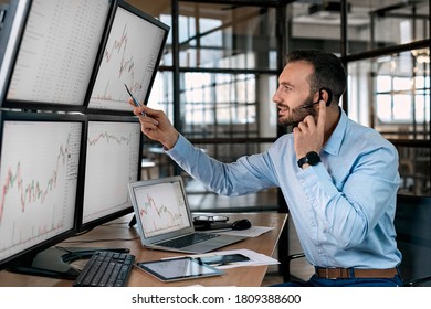 Bearded man trader wearing headset with microphone sitting at desk at office in front of monitors with data candle chart doing online trading training talking to employee explaining supply and demand