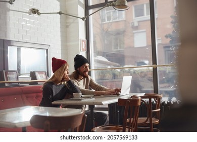 Bearded Man Typing On Laptop Computer While His Girlfriend Looking At It. Concept Of Romantic Relationship. Comfortable Lifestyle. Young European Couple Sitting At Table In Modern Cafe