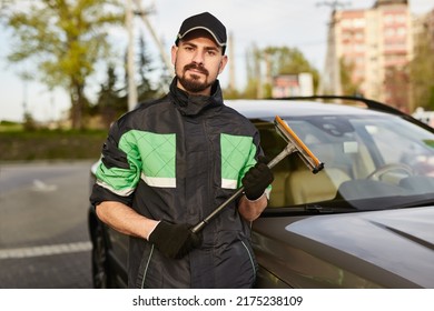 Bearded Man In Uniform And Cap With Sponge Looking At Camera While Standing Near Vehicle During Work At Gas Station In City