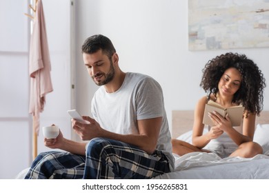 bearded man using smartphone while holding cup and sitting on bed near african american girlfriend reading book on blurred background - Powered by Shutterstock