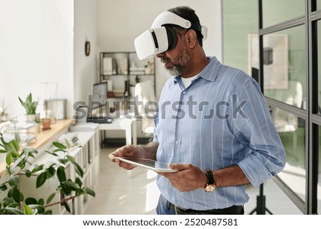 Bearded man using virtual reality headset while holding tablet in airy office with plants and large windows, deeply engaged with interactive virtual content