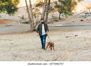 Bearded Man Walking His Dog Outside At Sunset. 40 Year Old Guy Walking With His Dog In The Countryside. Horizontal Photo With Copy Space