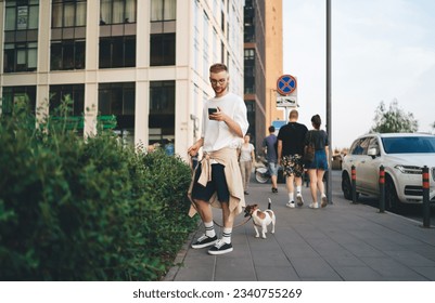 Bearded man wearing casual clothes and eyeglasses standing on sidewalk with Jack Russell Terrier dog on leash and reading message on social media on smartphone - Powered by Shutterstock