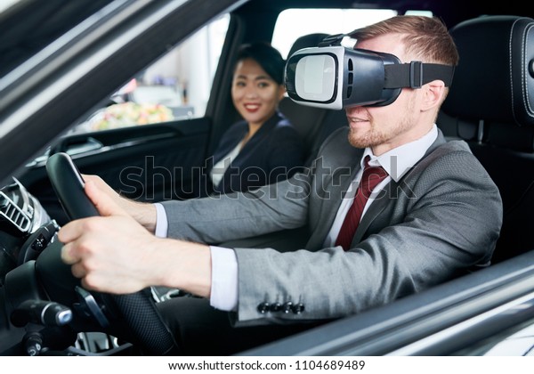 Bearded man\
wearing classical suit using VR headset while learning to drive\
car, pretty Asian instructor assisting\
him
