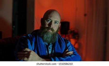 Bearded Man Wearing A Dressing Gown. Keeping Warm For The Winter. Warm Colour Background With The Man Wearing Cooler Colours. Arms Folded. Staring With Kind But Disappointed Eyes. 