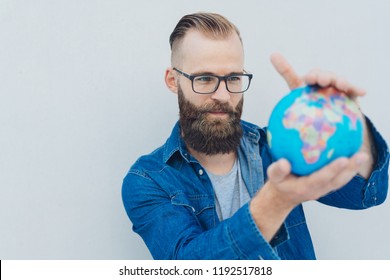 Bearded man wearing glasses holding up a world globe studying it with a look of concentration in a conceptual image of travel or conservation