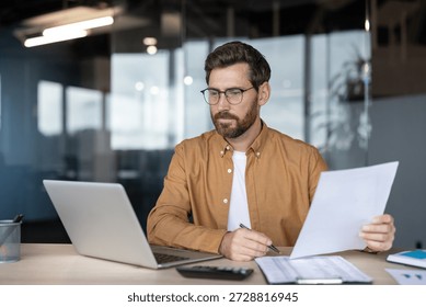 Bearded man wearing glasses processing data, reviewing financial reports, and working with documents and a laptop in a modern corporate office, focusing on productivity and planning