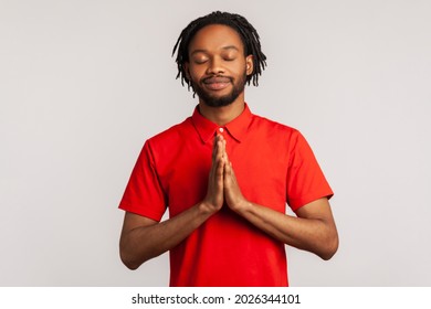 Bearded Man Wearing Red T-shirt, Concentrating His Mind, Keeping Hands Namaste Gesture, Meditating, Yoga Exercise Breath Technique Reduce Stress. Indoor Studio Shot Isolated On Gray Background.