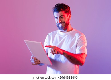 A bearded man wearing a white t-shirt interacts with a digital tablet while standing in a studio with purple and pink neon lighting. He is smiling and appears engaged with the content on the tablet. - Powered by Shutterstock