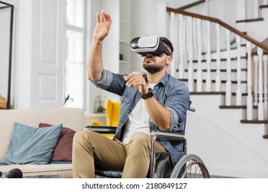 Bearded Man In Wheelchair Making Scrolling Gestures On Imaginary Screen Using Virtual Reality Headset. Caucasian Male Person With Special Needs Playing Favorite Game At Home.
