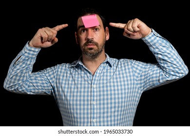 Bearded Man In A White And Blue Plaid Shirt Holding A Paper Stuck To His Forehead Pointing His Head With His Fingers, Isolated On Black Studio Background. Fun Concept