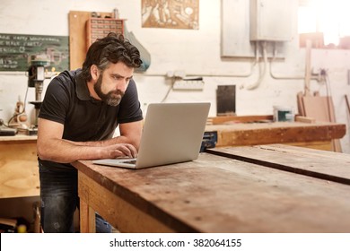 Bearded man who owns a small business, bending over at his work bench to type on his laptop, while working in his workshop and design studio - Powered by Shutterstock