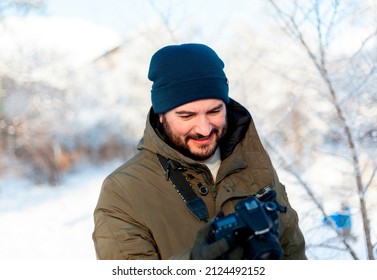 A bearded man in a winter hat and clothes holds a camera in mittens, looking at his screen. Photographer during photo shoot in winter outdoors