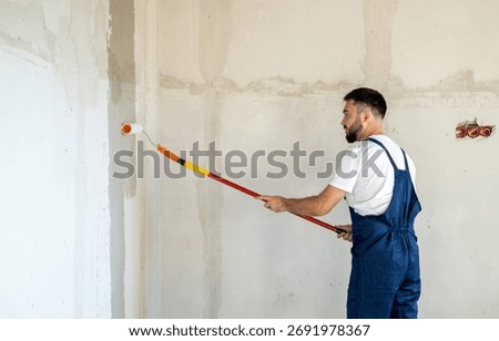A bearded man in a work uniform is priming the walls after applying gypsum plaster