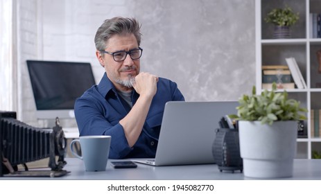 Bearded man working online with laptop computer at home sitting at desk. Businessman in home office, browsing internet. Portrait of mature age, middle age, mid adult man in 50s.