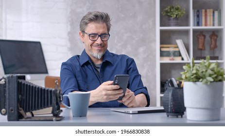 Bearded man working online with laptop computer and smart phone at home sitting at desk. Businessman in home office. Portrait of mature age, middle age, mid adult man in 50s.