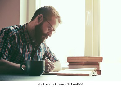 Bearded Man Writing With Pen And Reading Books At Table