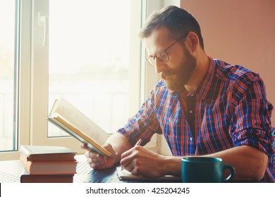 Bearded Man Writing With Pen And Reading Books At Table