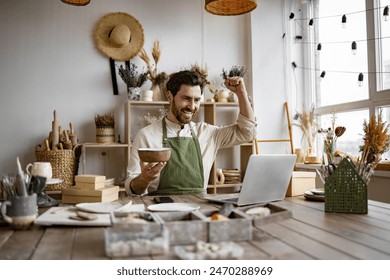 Bearded mature businessman engaged in retail trade of handmade tableware. Caucasian man potter happy with online sale of bowl on Internet raising hands up against background of shelves with ceramics. - Powered by Shutterstock