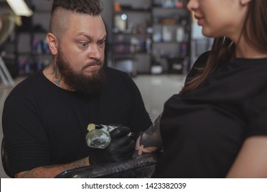 Bearded Mature Tattoo Artist Concentrating, Making Funny Face While Working On A Tattoo On A Female Client. Professional Tattooer Focusing With A Funny Face, Tattooing Young Woman