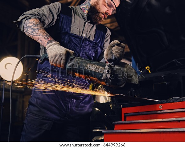 Bearded mechanic cuts steel car part with an\
angle grinder in a\
garage.