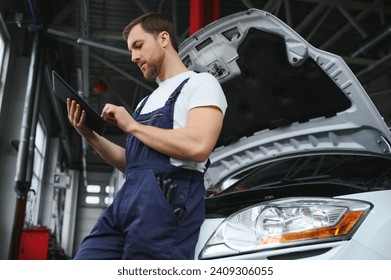 Bearded mechanic in overalls standing in garage of a car salon and holding tablet. He is about to diagnostic breakdown - Powered by Shutterstock