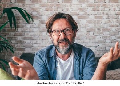 Bearded Middle Aged Man In Casual Clothes And Glasses Gesticulating And Talking To Camera While Sitting On Sofa Against Brick Wall In Daytime At Home