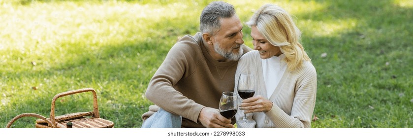 bearded middle aged man clinking glasses of wine with blonde and cheerful wife during picnic, banner