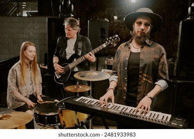 Bearded musician wearing hat and eyeglasses looking at camera while playing synthesizer, drummer and bassist discussing new song in background - Powered by Shutterstock