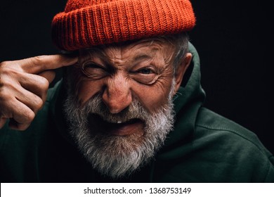 Bearded old man wearing orange knitted hat, looking at camera with crazy annoyed aggressive face expression making shooting gun gesture because tired of misery and insanity of life
