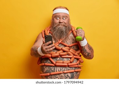 Bearded plump man stares at smartphone, reads something in internet, raises dumbbell, leads active lifestyle, wrapped with sausages around big tattooed belly, stands indoor, yellow background