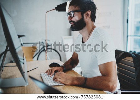 Bearded professional architect wearing eye glasses and white tshirt working at modern loft studio-office with desktop computer.Blurred background. Horizontal