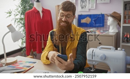 A bearded redhead man smiles while holding a smartphone in a colorful tailor shop, surrounded by sewing equipment and fabric swatches.