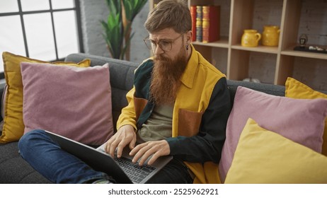 Bearded redhead man working on laptop indoors on colorful sofa surrounded by pillows and plants - Powered by Shutterstock