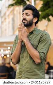 Bearded religion indian man praying with closed eyes to God asking for blessing, help, forgiveness outdoor. Adult guy clasping hands wishing luck in urban sunny city street. Town lifestyle. Vertical
